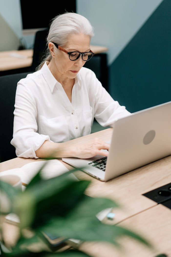 Senior woman with white hair using a laptop in a contemporary office setting, demonstrating focus and professionalism.